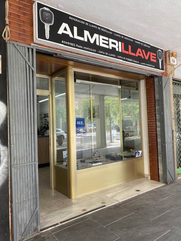 Locksmith storefront with a large sign that reads 'Reparación de llaves de vehículos' and 'ALMER LLAVE', glass display window showing keys inside.
