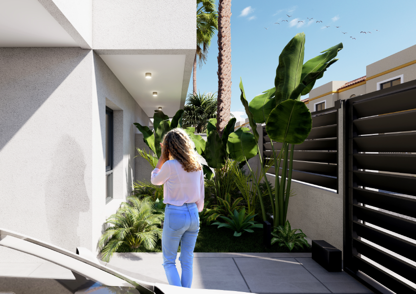 Oasis-Los-Pinos-II-Inalgra-Huercal-de-Almería-6 - MiraCasa Woman in a white blouse and blue jeans stands in a sunny modern courtyard filled with tropical plants