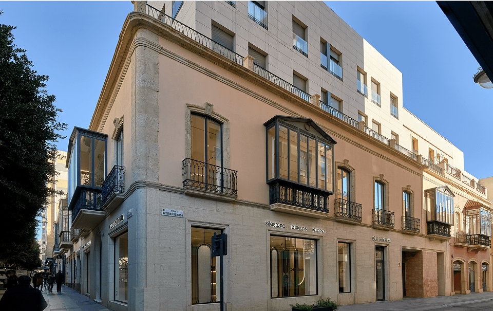 Corner of a beige historic building with wrought-iron balconies on a sunny city street; a modern structure rises behind it.